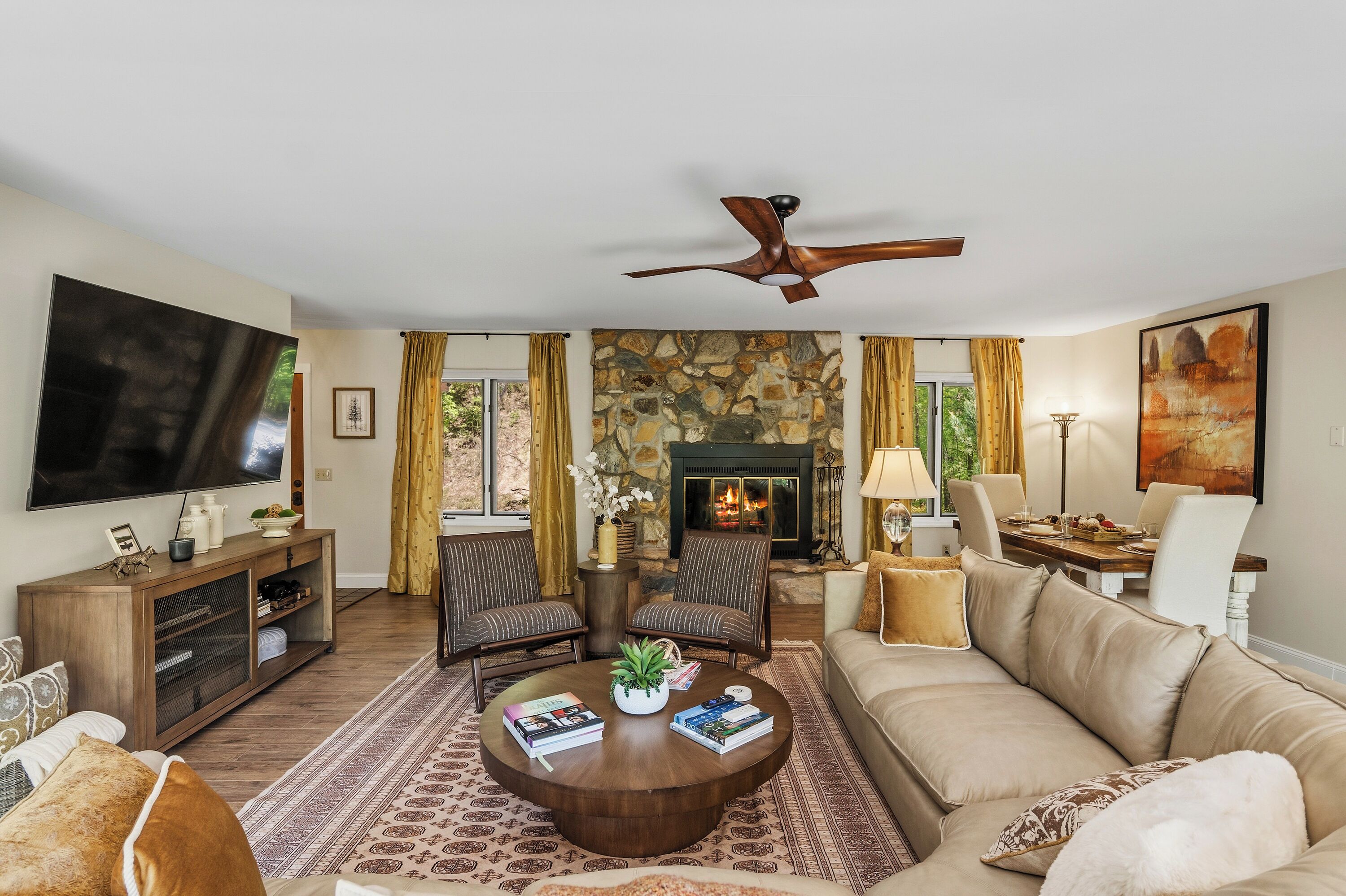 A living room with a stone fireplace, a flat-screen TV, a ceiling fan, and a dining area with chairs and a table.