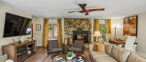 A living room with a stone fireplace, a flat-screen TV, a ceiling fan, and a dining area with chairs and a table.