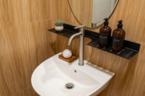 Un baño con un lavabo blanco, un espejo, una planta en maceta y dos botellas de jabón de manos y crema hidratante sobre un estante negro.