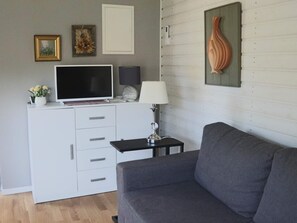 A modern living room with a grey sofa, a flat-screen TV on a white cabinet, a small table with a lamp, and a decorative vase on the wall. - 2 Person Holiday Home in Mellbystrand (Mellbystrand)
