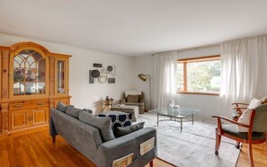 Ein Wohnzimmer mit Sofa, Sessel, Glastisch, Holzschrank und einem Fenster mit Vorhängen.