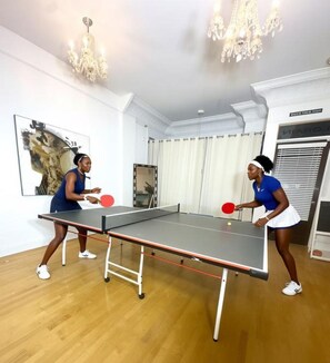 Zwei Personen spielen Tischtennis in einem Raum mit einem Tischtennistisch, Holzboden und einem Chandelier.