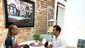 Dues persones jugant a bitlles en una habitació amb paret de maó, un quadre amb marc i una cadira blanca.