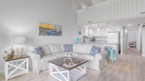 A spacious living area with a white sofa, a wooden coffee table, and a painting of a sunset over water. - Suite + Loft, Pool & Stunning Ocean Views! (Surf City)