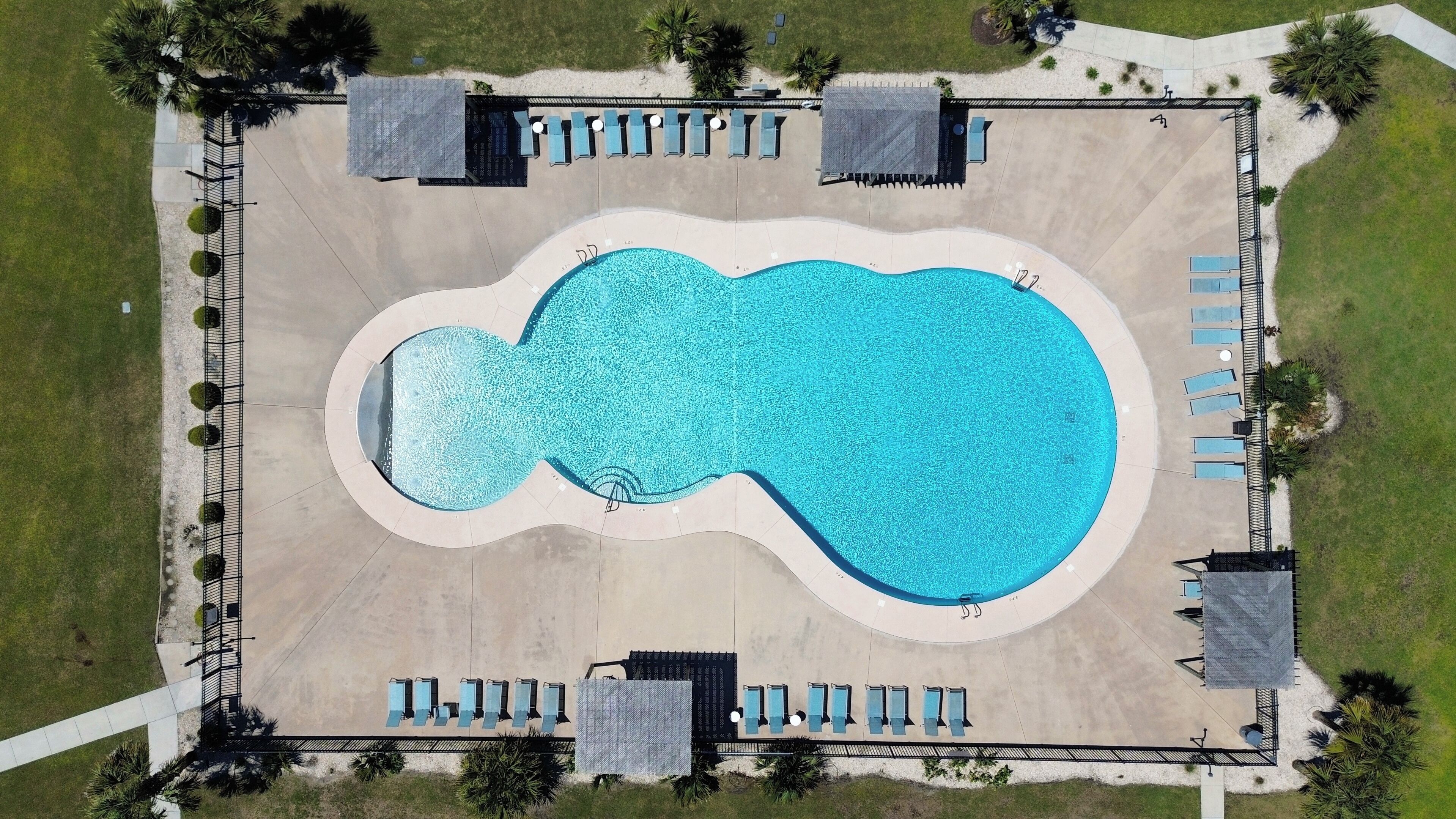 An aerial view of a swimming pool with a wavy edge, surrounded by lounge chairs and umbrellas, with a paved walkway and greenery in the background.