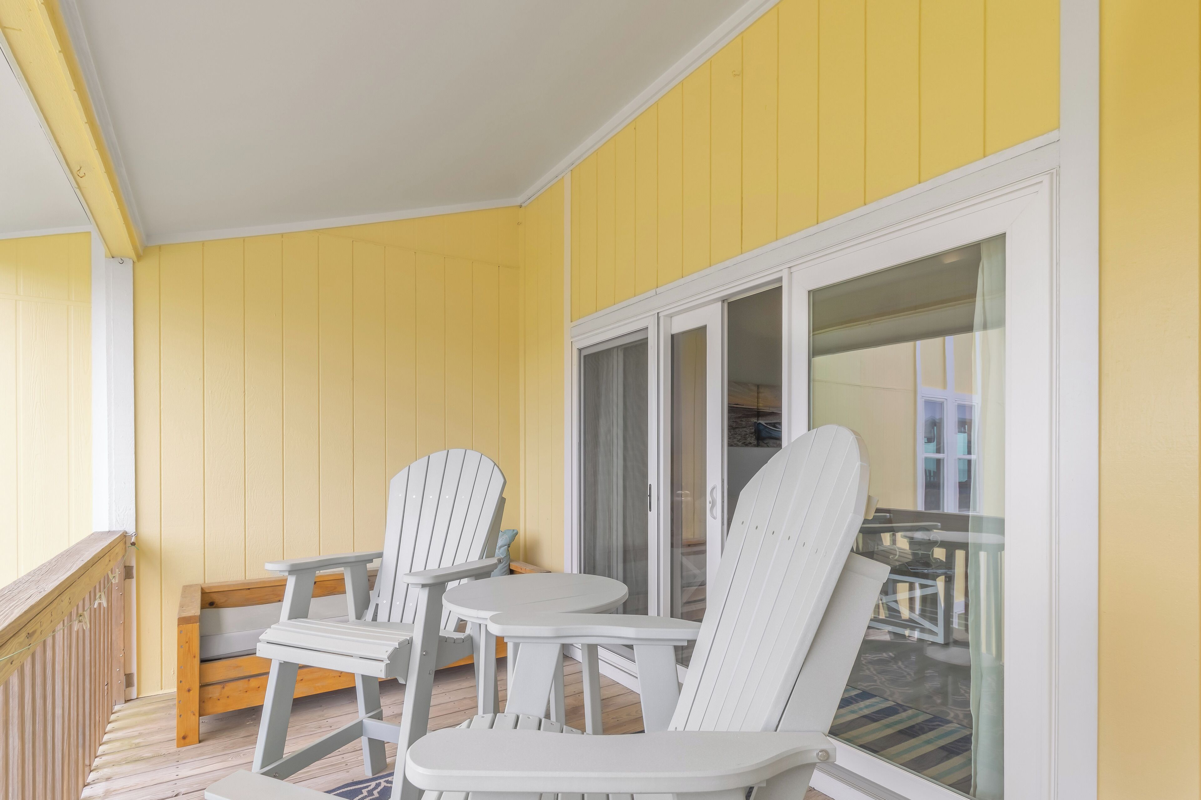 Un mur extérieur jaune, des portes vitrées à cadre blanc et deux chaises Adirondack blanches accompagnées d’une petite table.