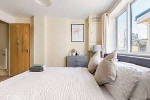 Ein Schlafzimmer mit einem Bett, einer Holztür, einem grauen Nachttisch, einem gerahmten Bild und einem Fenster.
