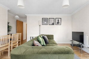 Ein Wohnzimmer mit einem grünen Sofa, Holzstühlen, einem Fernseher und gerahmten Kunstwerken an der Wand.