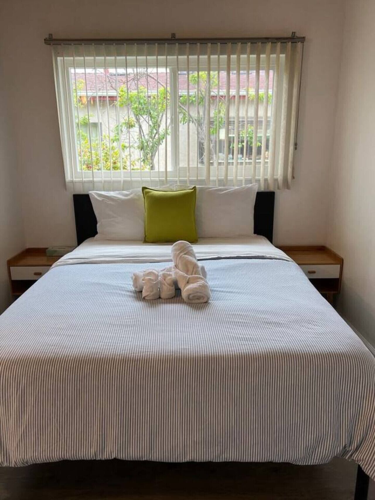 A hotel room with a bed, a window with blinds, a green pillow, and a neatly made bed with towels.
