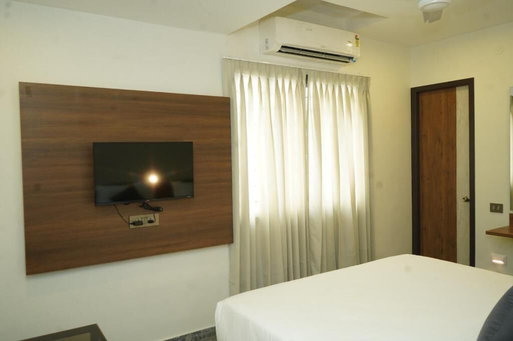 A hotel room with a bed, a mounted TV, a window with curtains, an air conditioning unit, and a door.