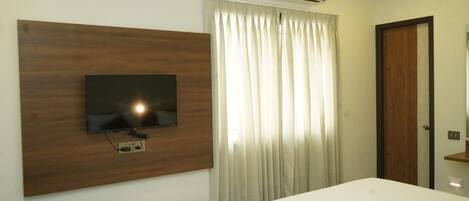 A hotel room with a bed, a mounted TV, a window with curtains, an air conditioning unit, and a door.