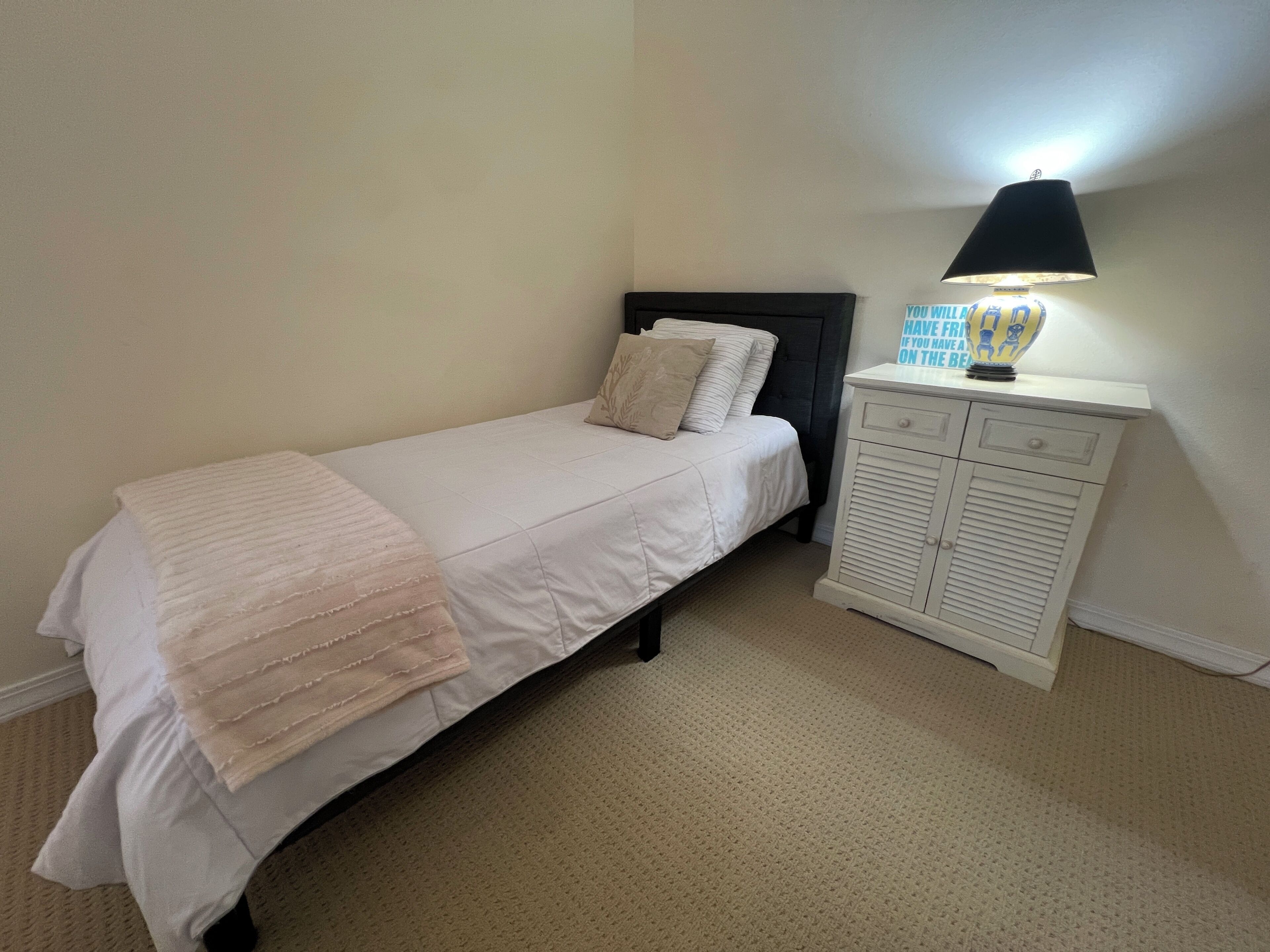 A single bed with a white bedspread and a beige throw blanket, a bedside table with a lamp, and a small cabinet with drawers.