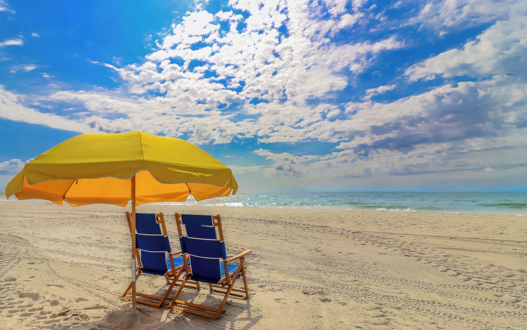 A beach scene with a yellow umbrella, two blue beach chairs, and a sandy beach under a partly cloudy sky.