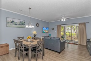 Un salon comprenant un canapé, une table à manger avec des chaises, un ventilateur de plafond et une vue sur l’extérieur, visible à travers une porte vitrée.