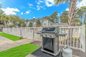 A barbecue grill with a gas tank and a propane cylinder, a white fence, and a well-maintained lawn. - The Coastal Catch Ground-Floor Oasis near Beach, Pool & Golf (Little River)