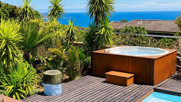 Un jacuzzi en una terraza de madera con piscina, rodeado de plantas tropicales y vistas a la costa.