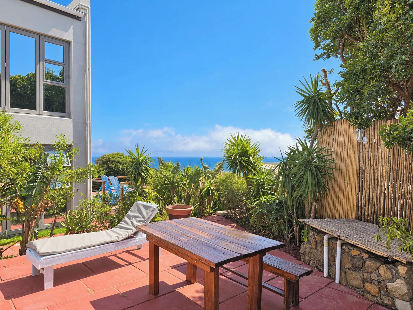 A patio with a wooden table and a lounge chair, surrounded by potted plants and a bamboo fence, overlooking a body of water.