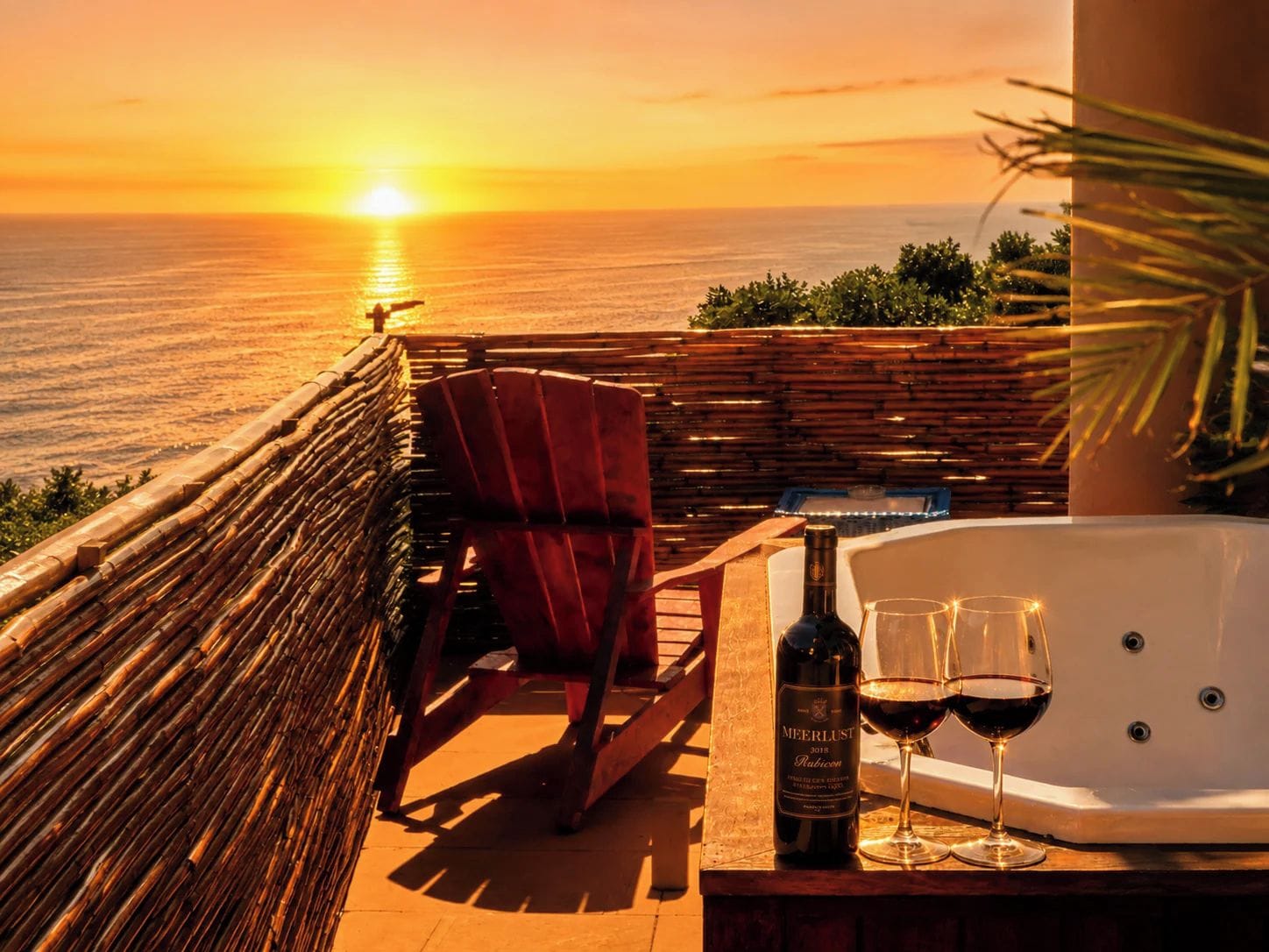 A sunset view from a balcony with a hot tub, a bottle of wine, and two glasses.