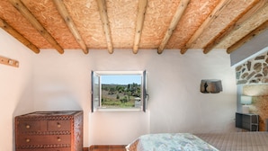 A bedroom with a wooden ceiling, a window with a view, a bed, a dresser, and a stone wall.