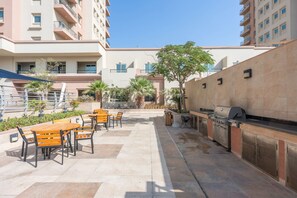 Eine Terrasse im Freien mit Holztischen und -stühlen, einem Grill und Blick auf ein mehrgeschossiges Gebäude.