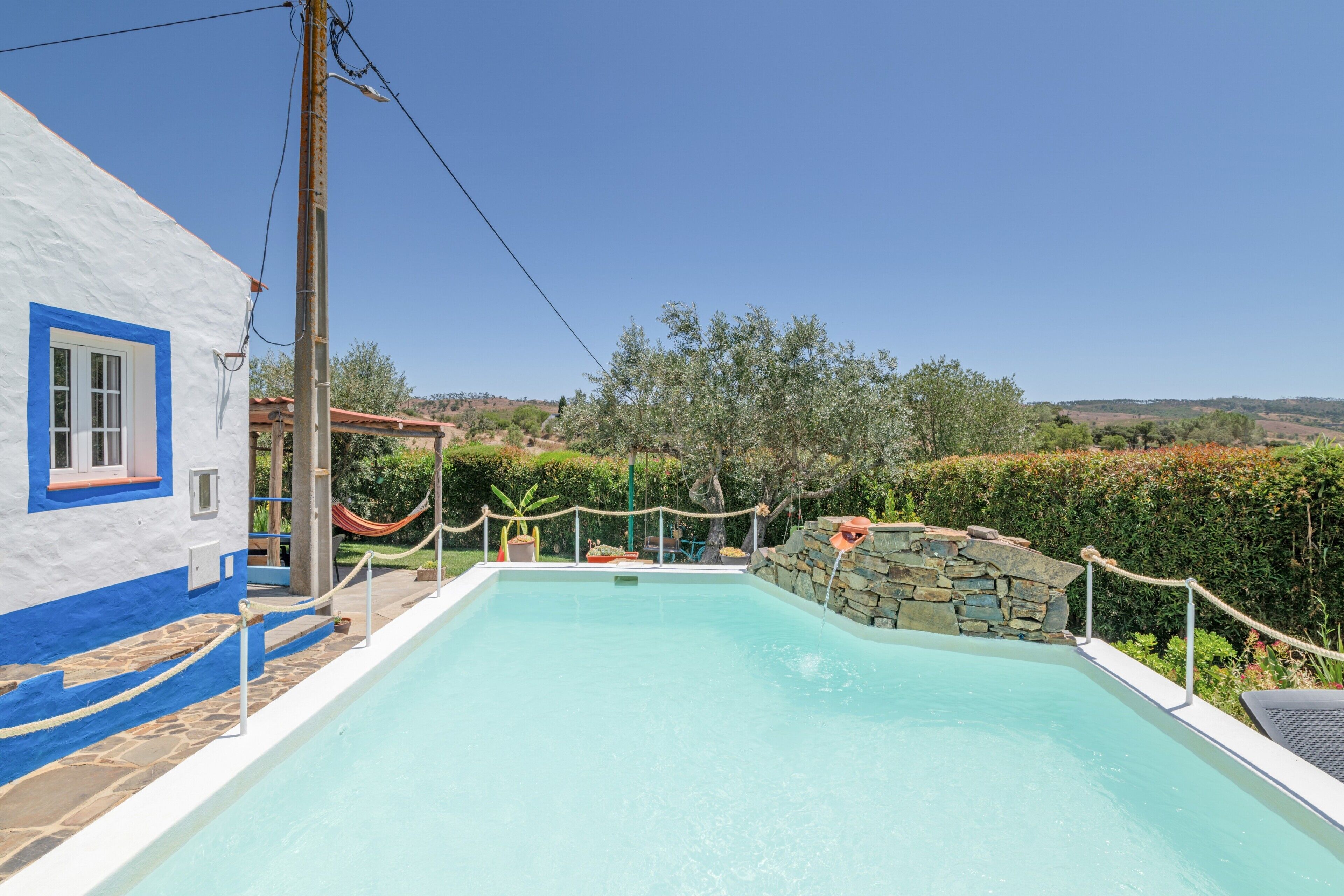 Una piscina con muro in pietra e barriera di corda, circondata da un edificio bianco con dettagli blu e vista sulla campagna.