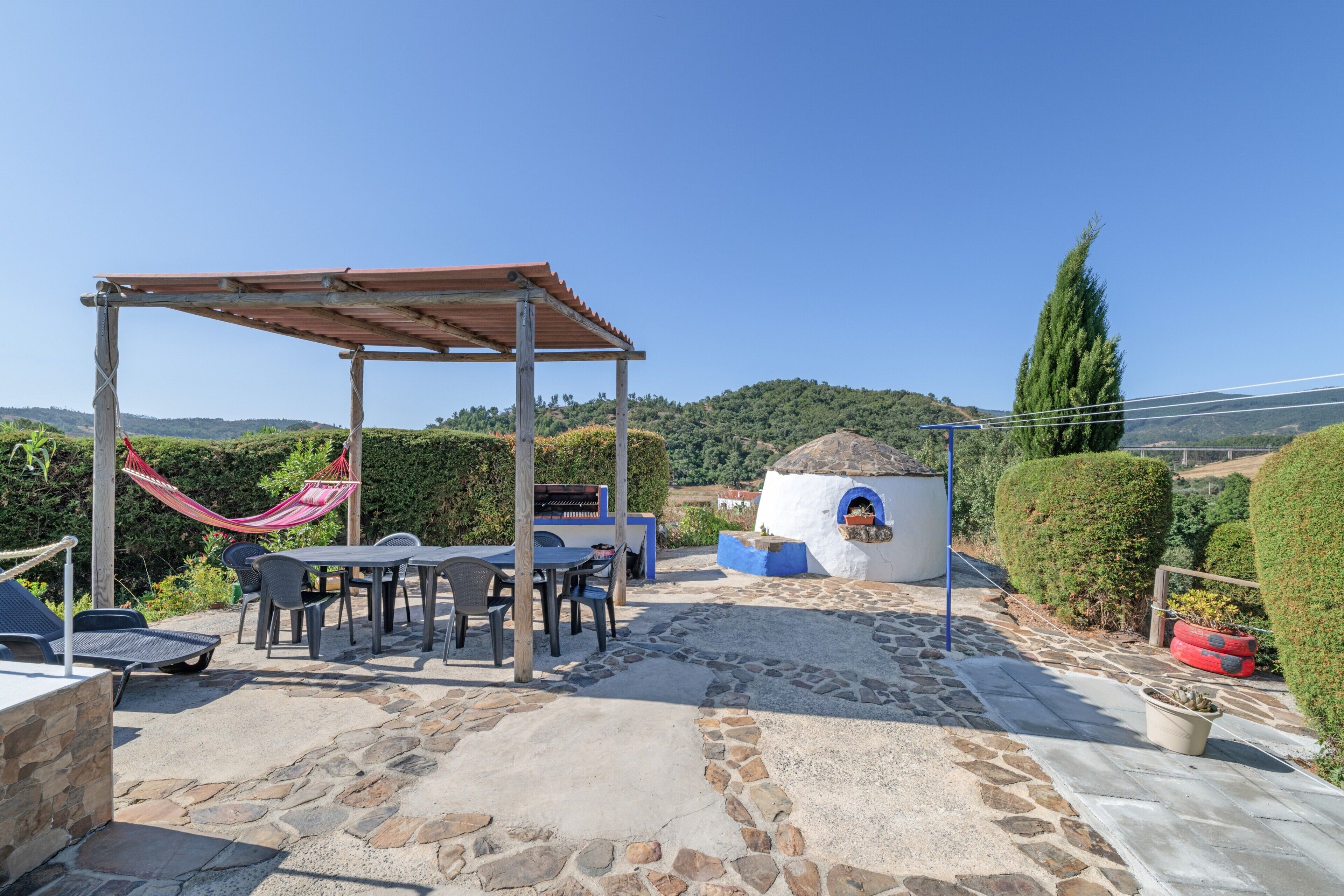 Een terras met stenen vloer, een houten pergola, een hangmat en een kleine buitenoven.