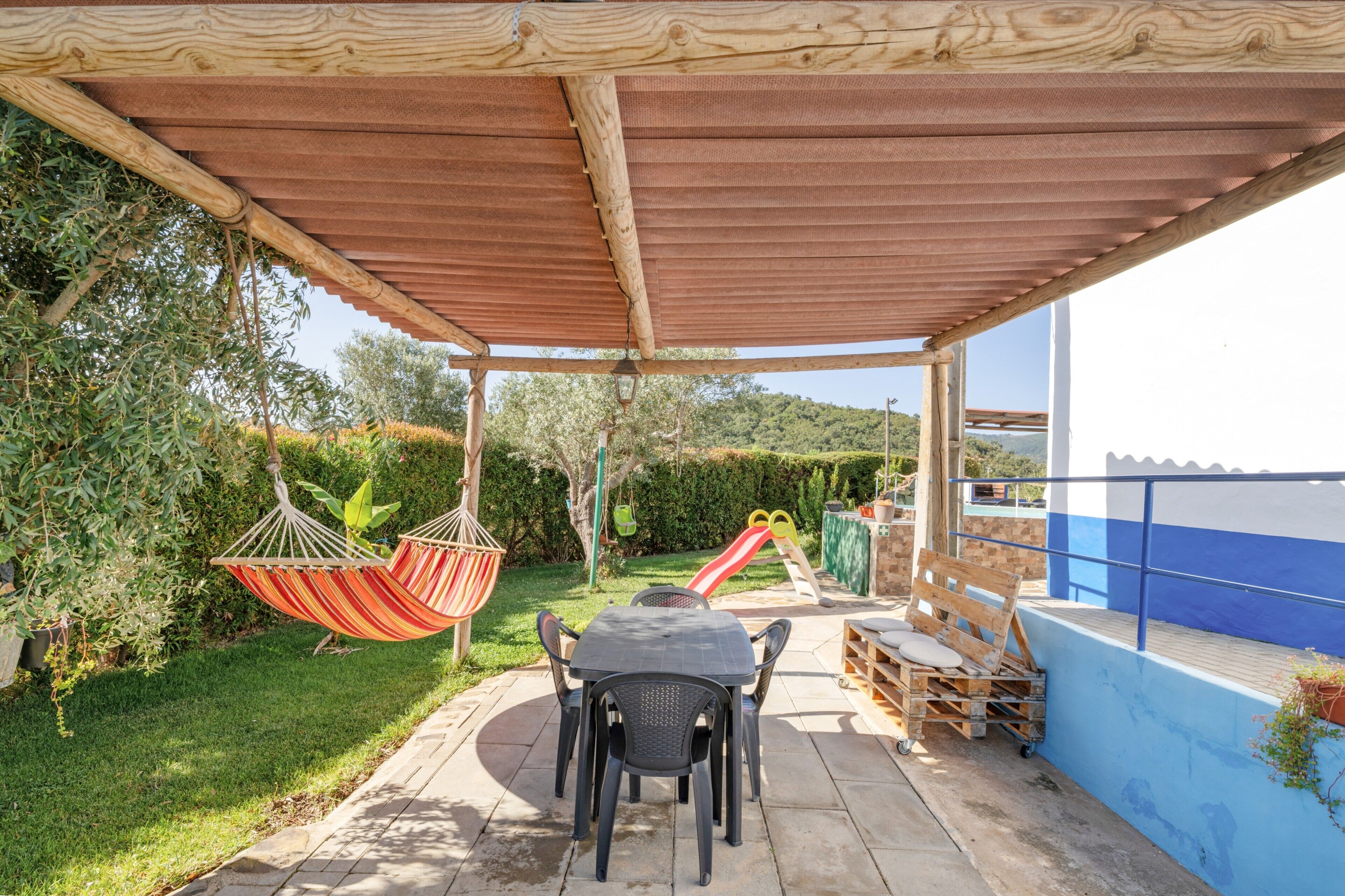 Eine überdachte Terrasse mit Tisch und Stühlen, einer Hängematte, einer Rutsche und Blick auf das Meer.