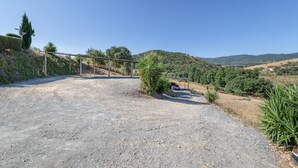 Ein befestigter Auffahrtsweg führt zu einem Metalltor, umgeben von Grünflächen und Hügeln im Hintergrund.