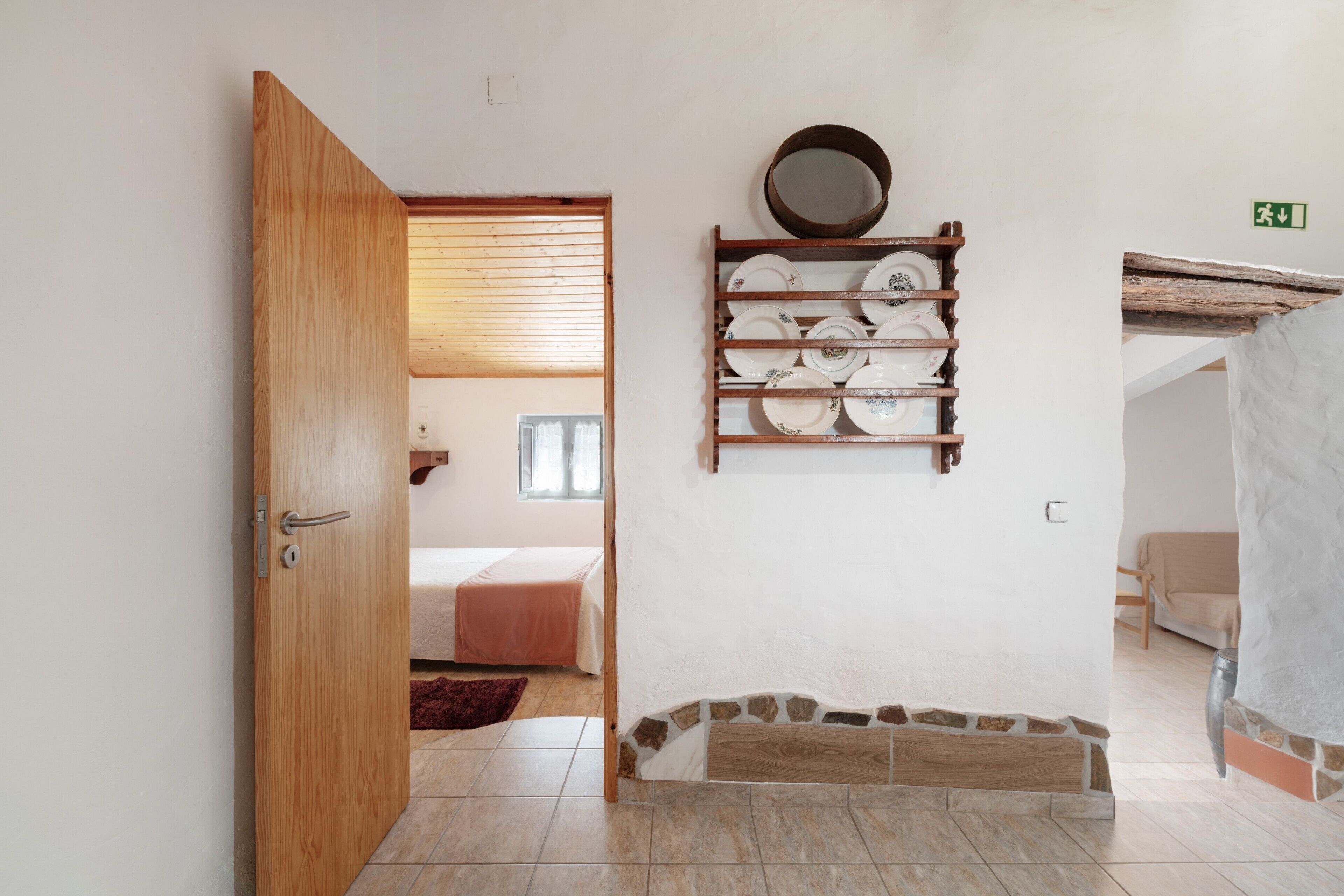 A room with a wooden door, a bed with a pink bedspread, a wall-mounted plate rack, and a stone accent wall.