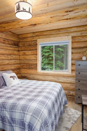 Un dormitorio tipo cabaña de troncos con una cama, un aparador, un ventanal con vistas a árboles y una lámpara con diseño de ciervo.