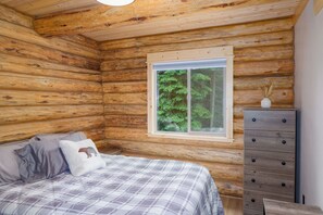 Ein Schlafzimmer in einer Blockhütte mit Bett, Kommode, Fenster und Blick auf Bäume.