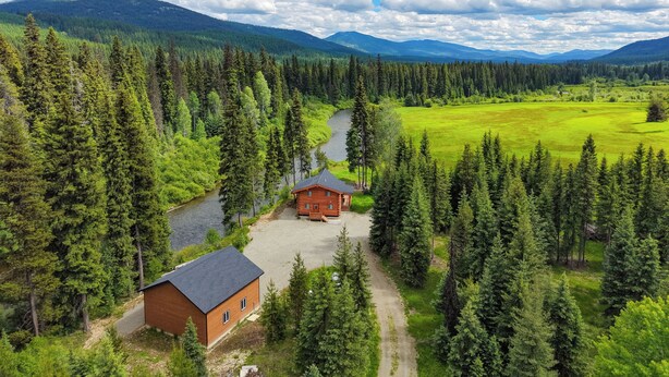 A cabin surrounded by dense forest, a river, and mountains in the background.