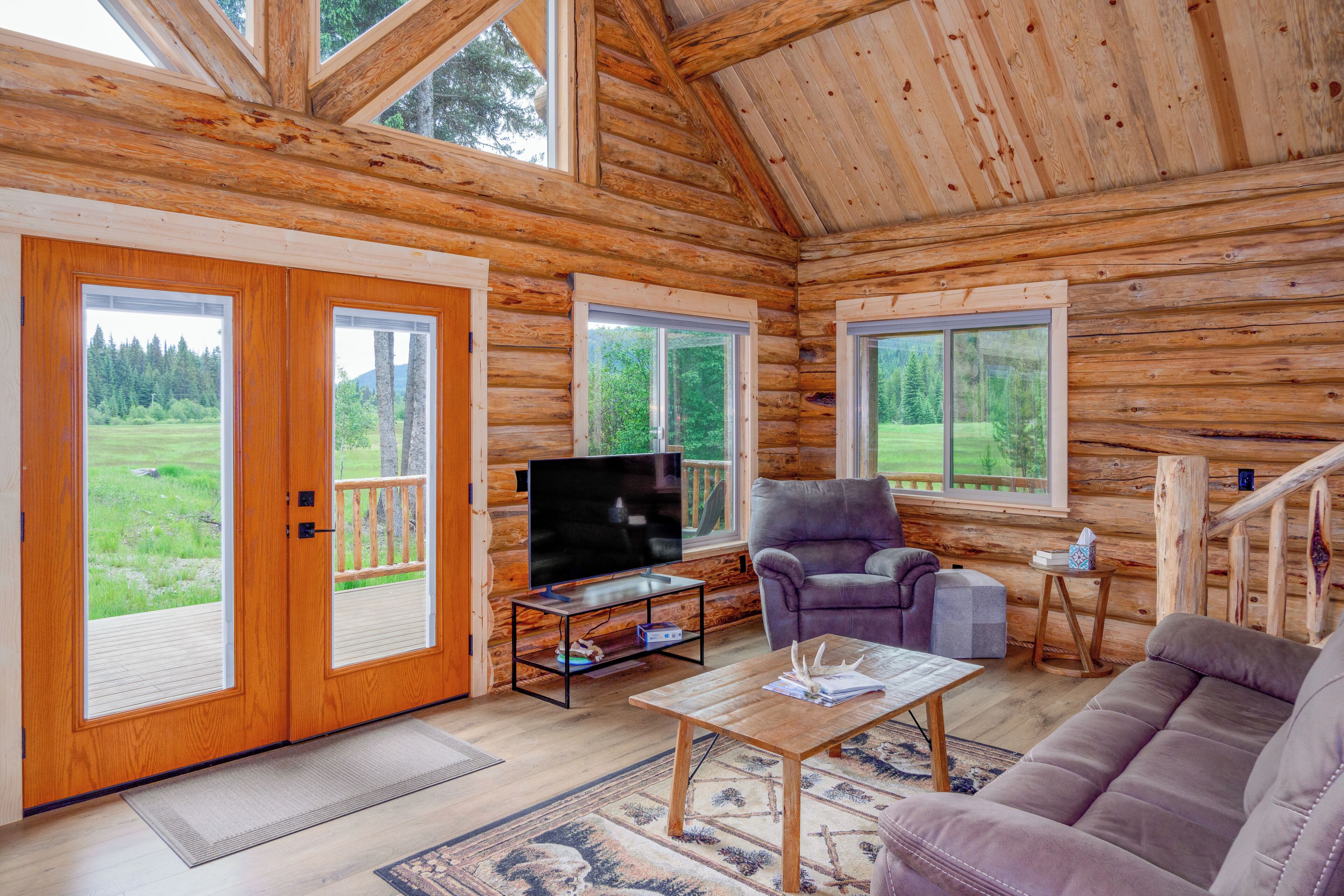L’intérieur d’une cabane en rondins, avec un téléviseur, un canapé et une vue sur un paysage verdoyant grâce à de grandes fenêtres.
