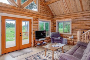 Ein Holzblockhaus-Interieur mit Fernseher, Sofa und Blick auf eine grüne Landschaft durch große Fenster.