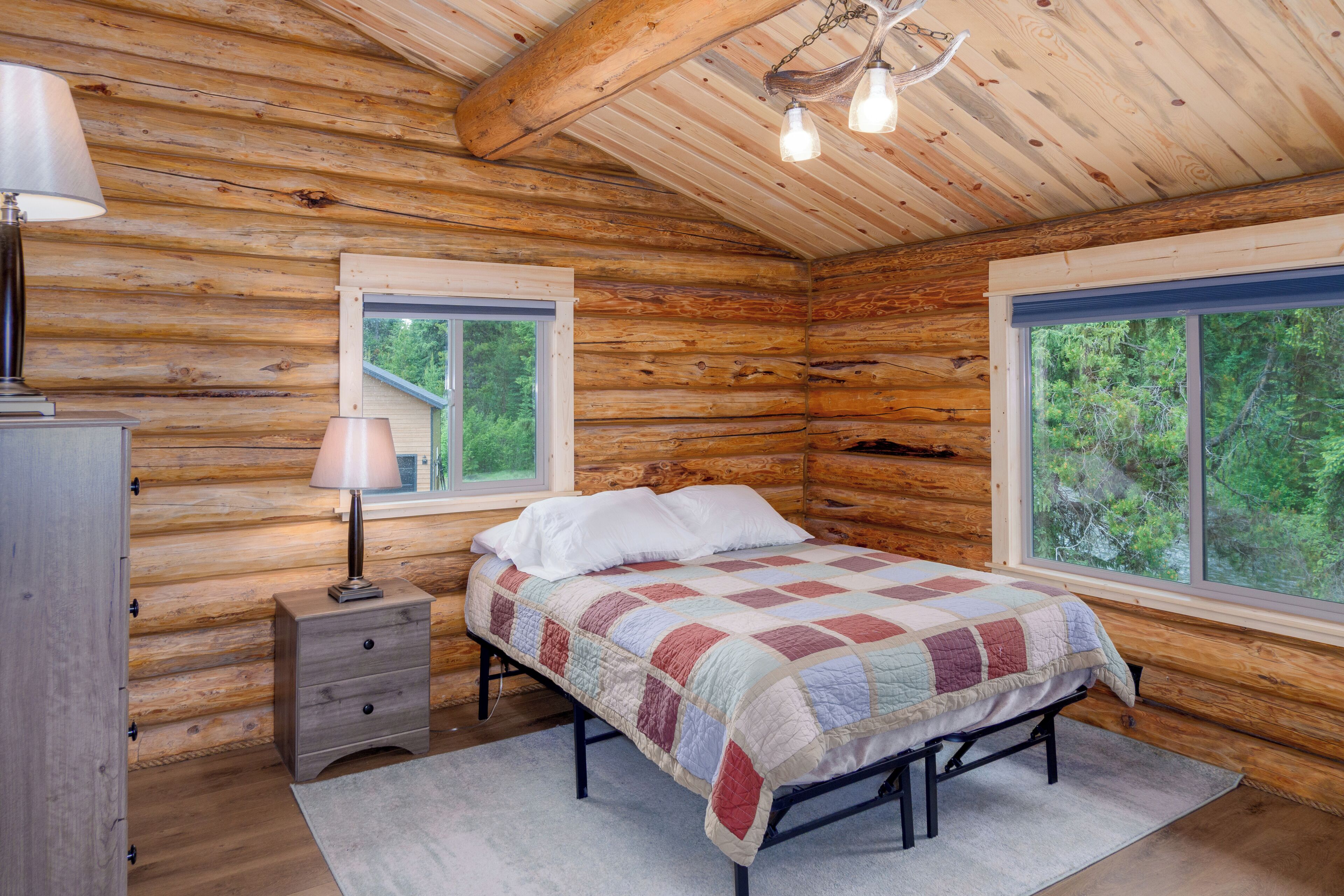 Une chambre à coucher située dans une cabane en rondins, avec un lit, deux fenêtres, une lampe et une commode.