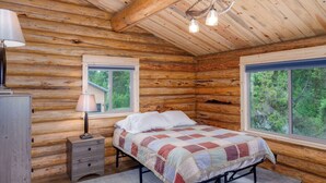 A log cabin bedroom with a bed, two windows, a lamp, and a dresser.
