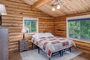A log cabin bedroom with a bed, two windows, a lamp, and a dresser.