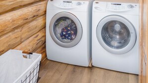 A log cabin interior with two white front-loading washing machines, a window, and a wooden floor.