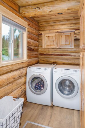 A log cabin interior with two white front-loading washing machines, a window, and a wooden floor.