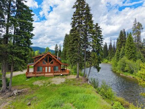 A log cabin with a deck, surrounded by a river and forest. - Escape to the Yaak - Your Montana Wilderness Retreat (Troy)