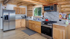 A log cabin kitchen with a stainless steel refrigerator, wooden cabinets, a microwave, a stove, and a large window.