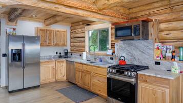 Une cuisine rustique aménagée dans une cabane en rondins, avec un réfrigérateur en acier inoxydable, des armoires en bois, un four à micro-ondes, une cuisinière et une grande fenêtre.
