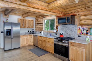 A log cabin kitchen with a stainless steel refrigerator, wooden cabinets, a microwave, a stove, and a large window.