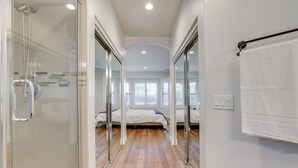 Une chambre d’hôtel moderne dotée d’une douche en verre, d’un plancher en bois, d’un lit avec une literie blanche et d’une grande fenêtre laissant entrer la lumière naturelle.