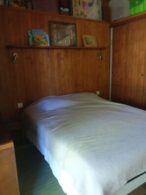 Een kamer met een bed, houten lambrisering, een plank met boeken en een lijst met een afbeelding, en een deur.