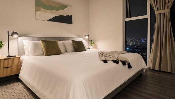 Una habitación de hotel moderna con una cama grande, lámparas de noche, vistas a la ciudad y un cuadro en la pared.