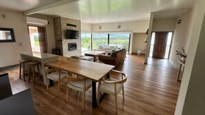Eine moderne Küche mit einer zentralen Kochinsel, ein Essbereich mit Holztisch und -stühlen, ein Wohnzimmer mit Sofa und Fernseher, sowie große Fenster mit Blick ins Freie.