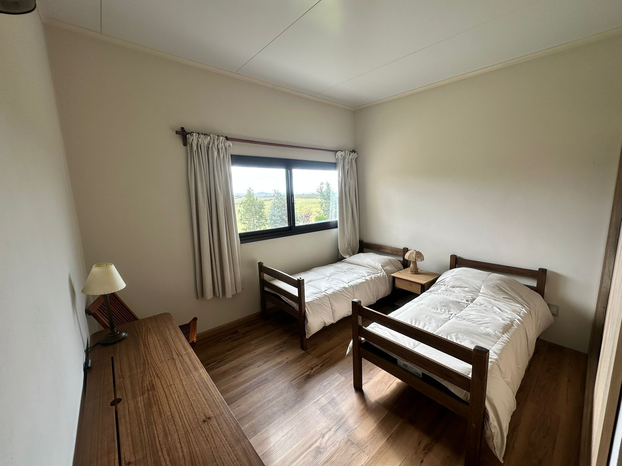 Une pièce comprenant deux lits en bois, une petite table avec une lampe, une fenêtre avec des rideaux et un bureau en bois.