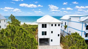 A white building with a garage, surrounded by pine trees, near a beach. - NEW- 6 bedroom-Private Pool- Pet Friendly (Saint George Island)