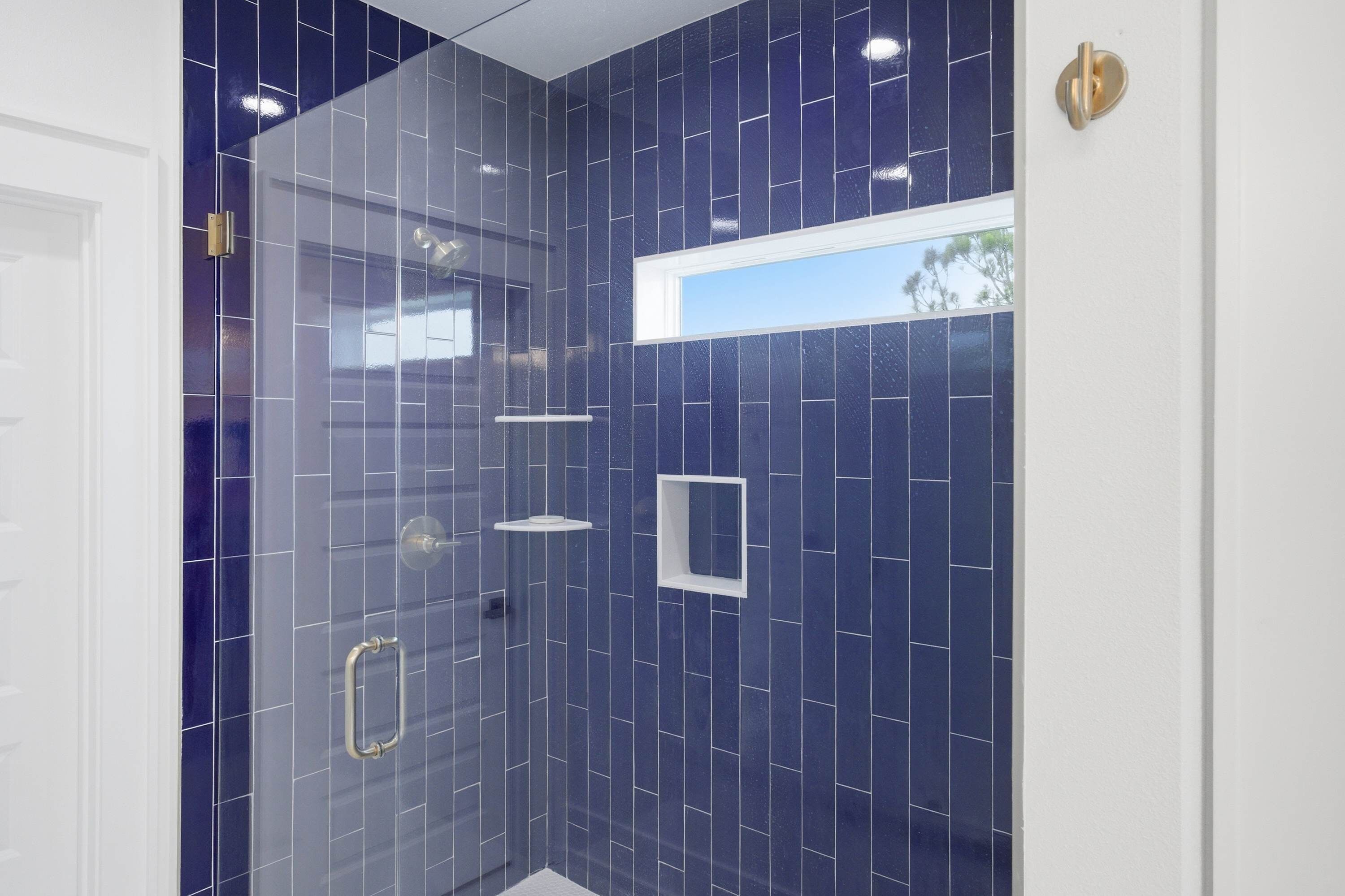 A modern bathroom with dark blue tiles, a glass shower enclosure, and a small window.